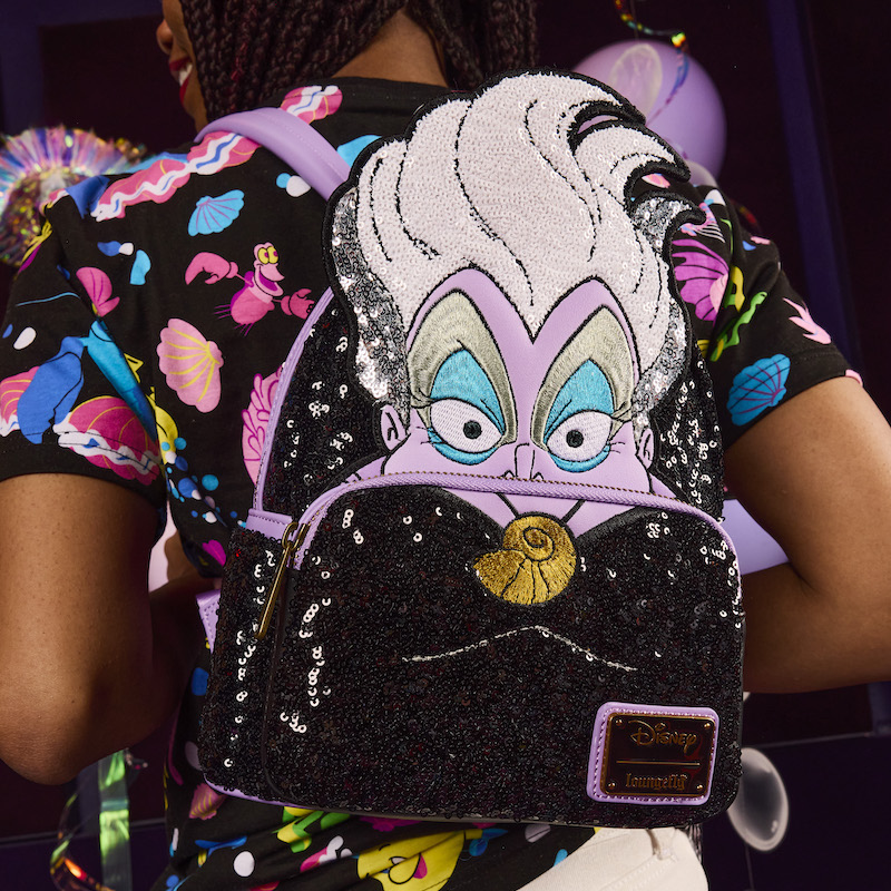 Woman wearing the Loungefly Little Mermaid 35th anniversary unisex tee and wearing the Exclusive Ursula Sequin Mini Backpack, featuring Ursula as a large appliqué with embroidered facial details and white and silver sequins on her hair. The rest of the bag is covered in black sequins. 